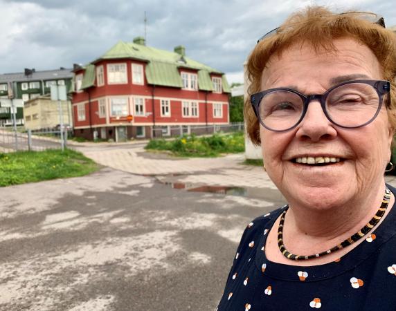 Majvor har kort brunt hår och bär glasögon. Hon ser glad ut och står framför ett hus i gamla Kiruna.