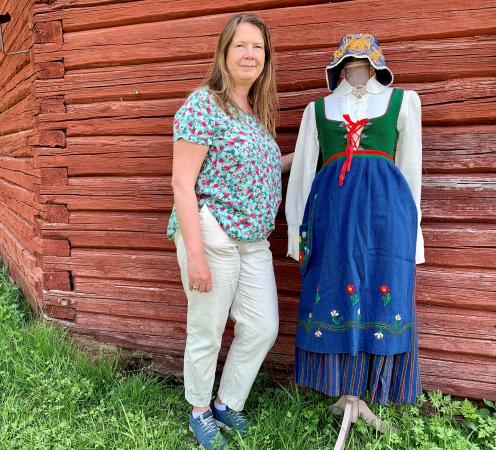 Anja står framför en röd timrad vägg bredvid 1912-års Norrbottensdräkt i blått, vitt och grönt.