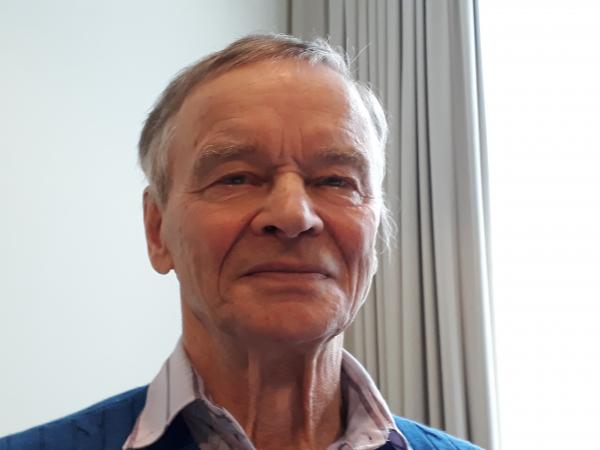Taisto har grått, kort hår och ser stolt ut på bilden. Han har en vit skjorta med en blå tröja över.