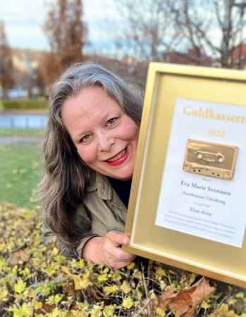 Eva Marie Svensson, som är redaktör för taltidningen Insyn, håller upp en gyllene tavla med en guldkassett i mitten. Hon lutar sig mot en höstklädd gräsmatta och ler. Läpparna är röda och håret utsläppt.