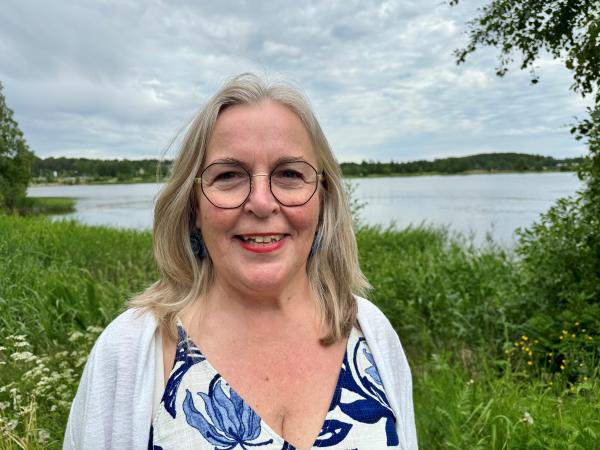 Anna-Maria har mellanblont hår till strax nedanför axlarna. Hon bär en blåmönstrad klänning och vit kofta. Anna-Maria har glasögon och ser glad ut. Hon står framför vatten och grön växtlighet.
