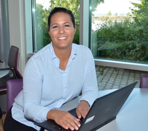 Kristina Larsson har mörkt hår i hästsvans, bruna ögon och en ljus skjorta som är öppen i halsen. Hon sitter vid ett bord och har en bärbar dator framför sig.