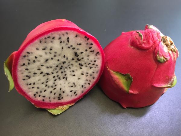 En uppskuren pitaya ligger på svart underlag. Skalet är rödlila och fruktköttet vitt med små svarta kärnor