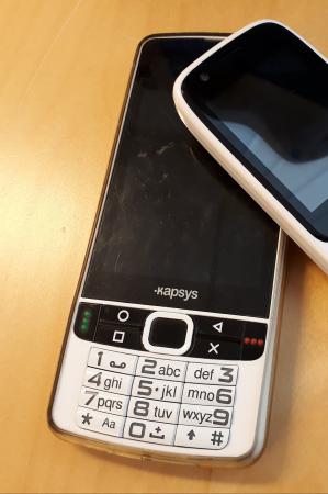 Telefonen har svart skärm och vit knappsats. Den ligger på ett bord.