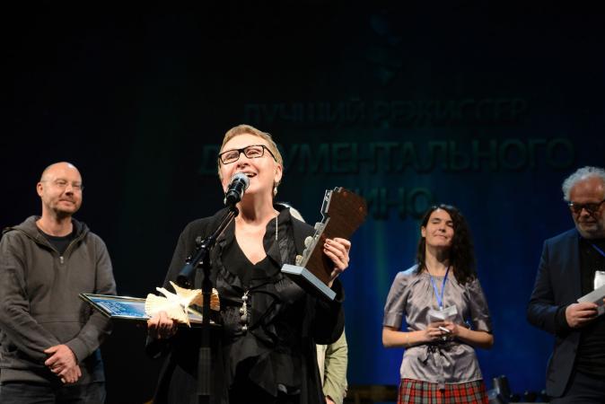 Foto: Maxim Churov Valentina ger ett tacktal på scenen i mikrofon. Hon har kort mörkt hår och glasögon med mörk båge. Hon ser glad ut och håller diplom och prisstatyett. bakom henne står tre av jurymedlemmarna som utsåg henne till årets regissör på filmfestivalen Arctic Open, Arkhangelsk, Ryssland.