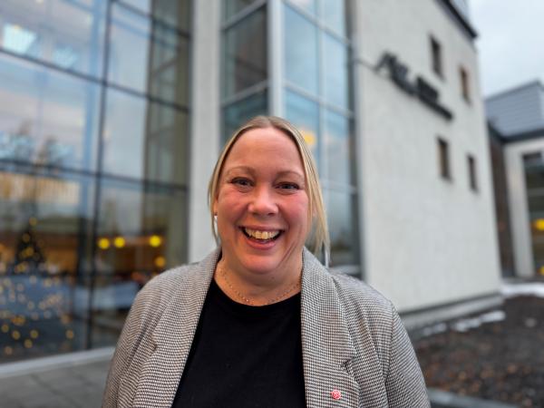 Camilla är blond och har håret uppsatt. Hon bär en grå tröja över en svart tunika. Hon står framför Regionhuset i Luleå och ser glad ut.