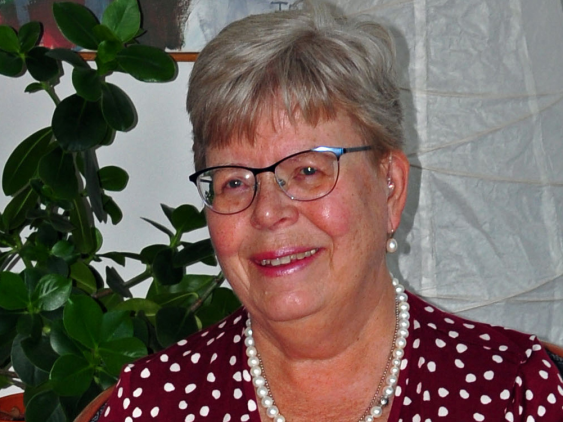 Marja Liisa har grått hår och bär glasögon. Hon bär en mönstrad blus och ett pärlhalsband. Hon ser glad ut.