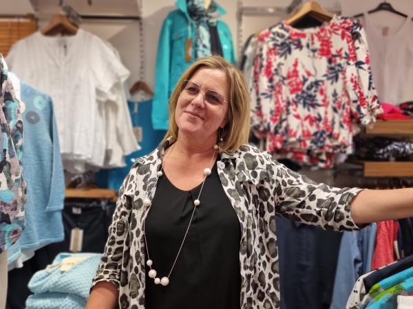 Malin Jönsson står i sin butik framför massa olika kläder som hänger i bakgrunden. Hon har en svart tröja, med en svartvit leopardmönstrad skjorta över. Hon har ett långt halsband med stora vita pärlor och stora glasögon. Håret är utsläppt och hon ler litegrann.