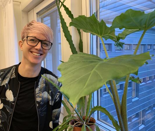 Malin har kort ljusrosa hår och mörka glasögon. Hon ser glad ut bland växter med stora blad