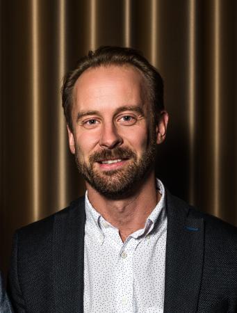Daniel Nilsson har brunt hår och skägg. Han har en vit skjorta med små mörkblå prickar. Utanpå har han en mörk kavaj.