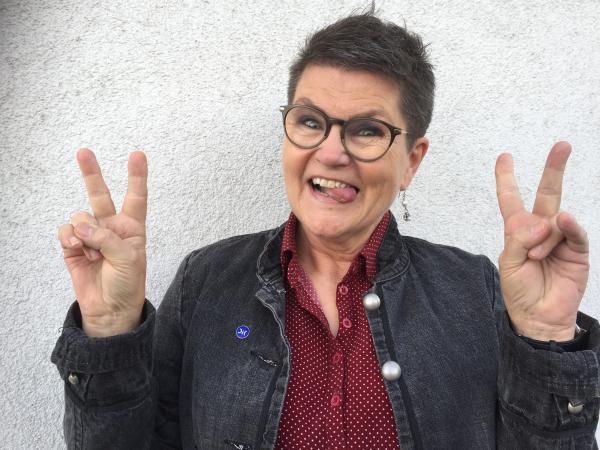 Agneta har kortklippt, mörkt hår och glasögon med mörka bågar. Hon har på sig en röd blus och en svart jeansjacka och hon gör v-tecknet med båda händerna.