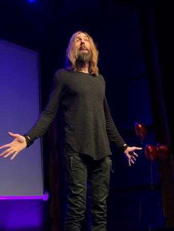 Micael är klädd i svart och har långt mellanbrunt hår och skägg. Han står på en scen med armarna utslagna och ser frågande ut.