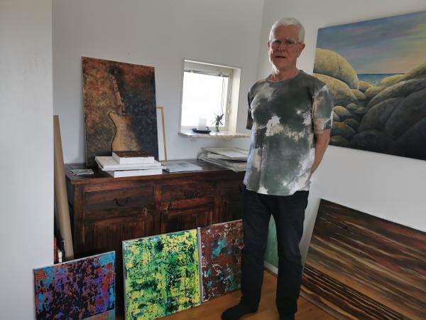 Staffan Björlin står i sin ateljé. Han har flera målningar intill sig, i starka färger. Det ser ut som abstrakt konst. I bakgrunden syns ett litet fönster med en ljusstake och en ängel. Han har en gråvit kortärmad tröja på sig och ett par svarta byxor. Han har glasögon och vitt kort hår.