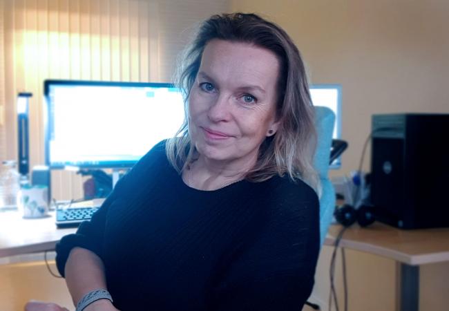 Maria har halvlångt mellanblont hår och blå ögon. Hon sitter på sitt kontor med dataskärmen bakom sig och bär en mörkblå stickad tröja. Hon ser glad ut!