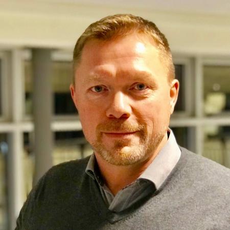 Rickard har kort rött hår och skägg. Han har en grå tröja med skjorta under.