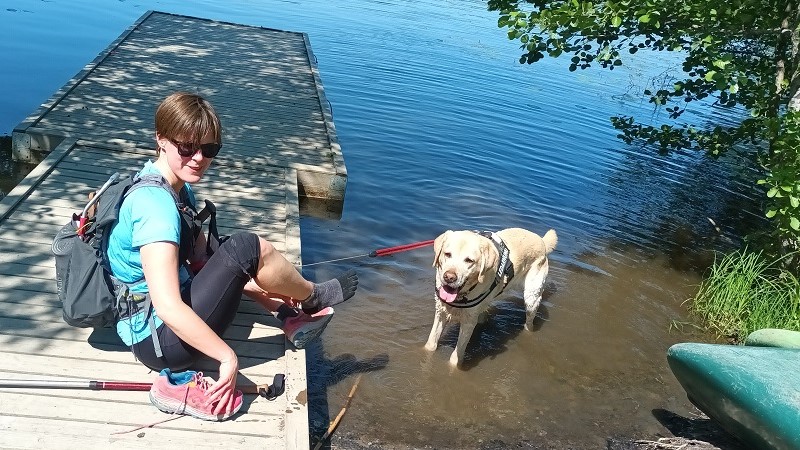 Ida sitter på en brygga och bär ryggsäck. Hennes ledarhund är i vattnet.