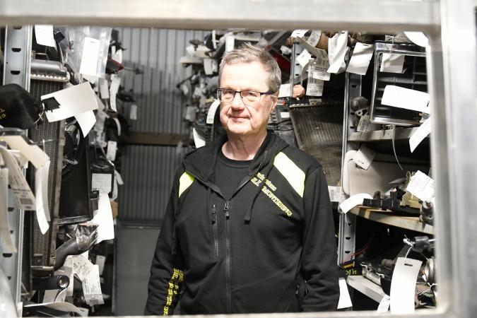 Ingvar står mitt i lagret med bildelar. Han har en svart jacka och bär glasögon