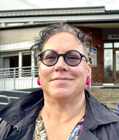 Greta har kort mörkt hår med ljusa slingor. Hon bär glasögon och rosa örhängen. Hon ser glad ut.