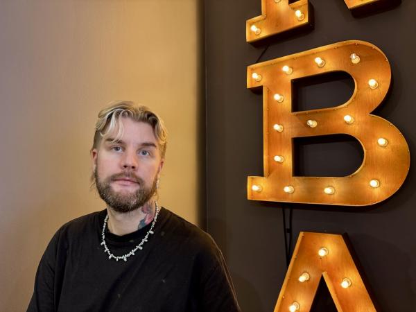 Andreas har gröna ögon och håret i en hockeyfrilla. Han har skägg och står vid sidan av bokstaven B med lampor i Frisör Andreas
