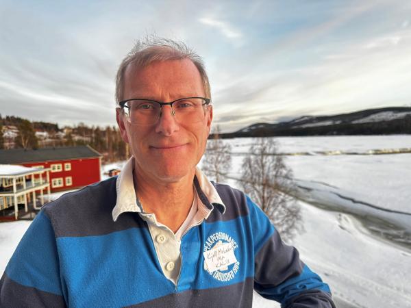 Kjell har kort hår och bär glasögon. Han har en blå randig tröja och står på en balkong med utsikt över Storforsen. Kjell Melander