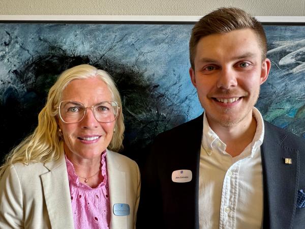 Ann Christine har långt blont hår och glasögon. Hon bär en rosa blus och en benfärgad kavaj. Jens har mörkt, kort hår och har en svart kavaj och vit skjorta. De står framför ett konstverk och ser glada ut. Ann Christine Robertson och Jens Erkheikki