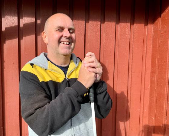 Robert har inget hår och är klädd i en Skellefteå AIK-tröja i svart, gult och grått. Han håller i sin käpp och ser glad ut.